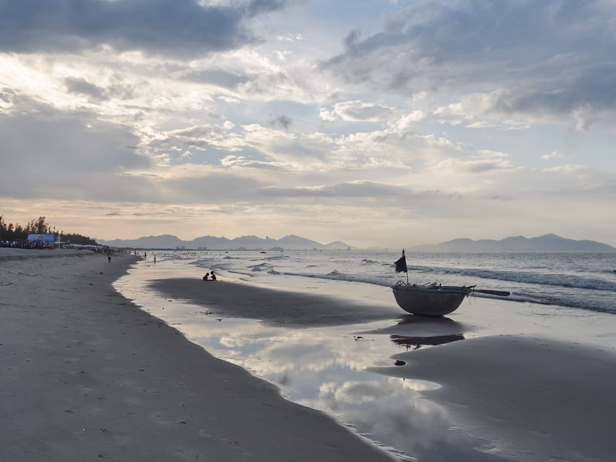 A wide sandy beach at low tide, a round basket boat at the water’s edge and blue mountains on the horizon