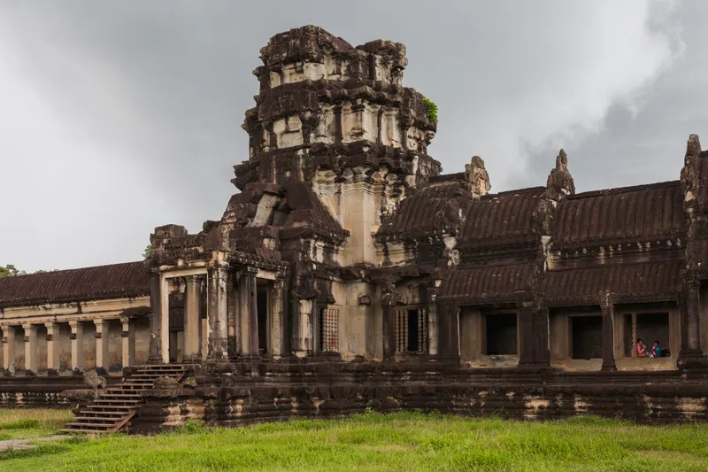 The weathered sandstone gate tower and long roofed galleries of Angkor Wat under heavy grey cloud, with bright green grass in front