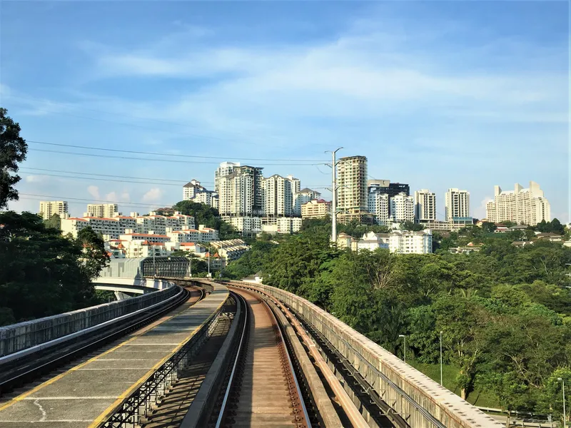A view along an elevated rail line towards a hill of white apartment blocks and low-rise houses among trees, under a blue sky