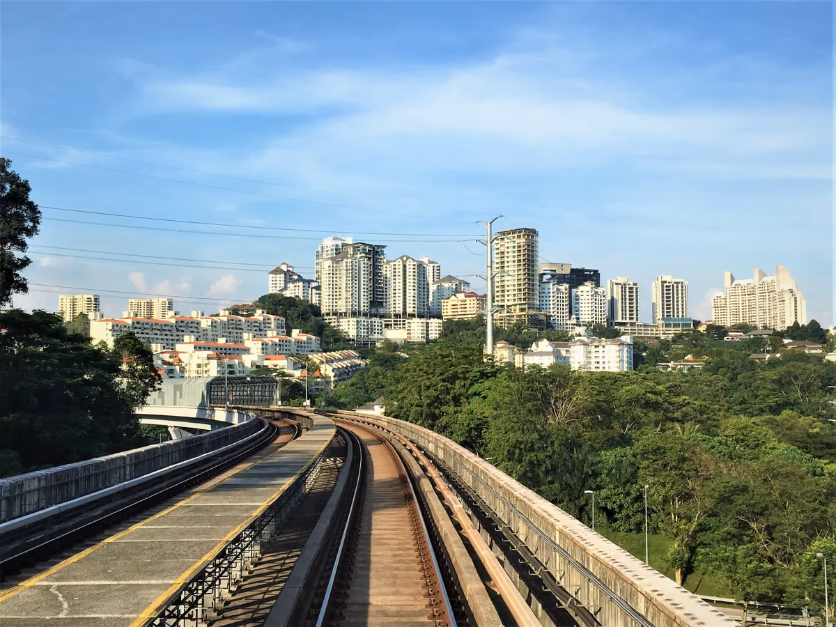 A view along an elevated rail line towards a hill of white apartment blocks and low-rise houses among trees, under a blue sky