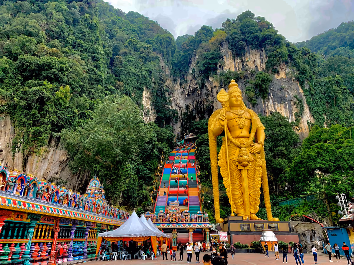 Steep stairs painted in bands of colour climb a forested limestone cliff, beside a giant golden statue of a Hindu deity