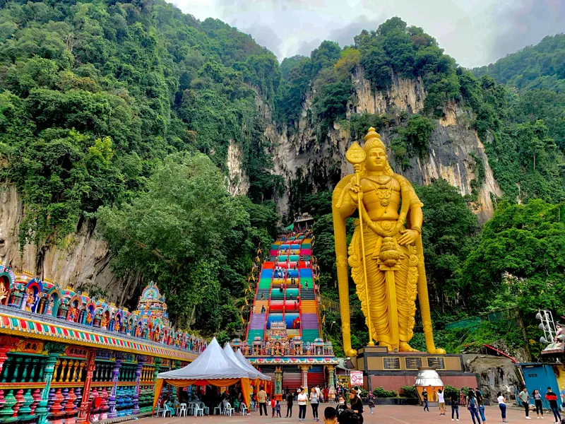 Steep stairs painted in bands of colour climb a forested limestone cliff, beside a giant golden statue of a Hindu deity