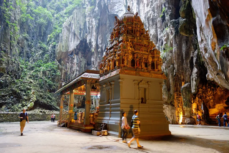A carved and painted Hindu shrine tower on the floor of a huge limestone cave, with sheer rock walls, plants growing on them, rising to daylight above