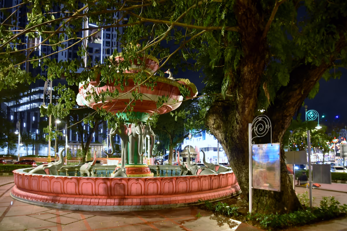 A tiered pink fountain on a base of carved elephants, lit at night under a large tree, with an apartment tower behind
