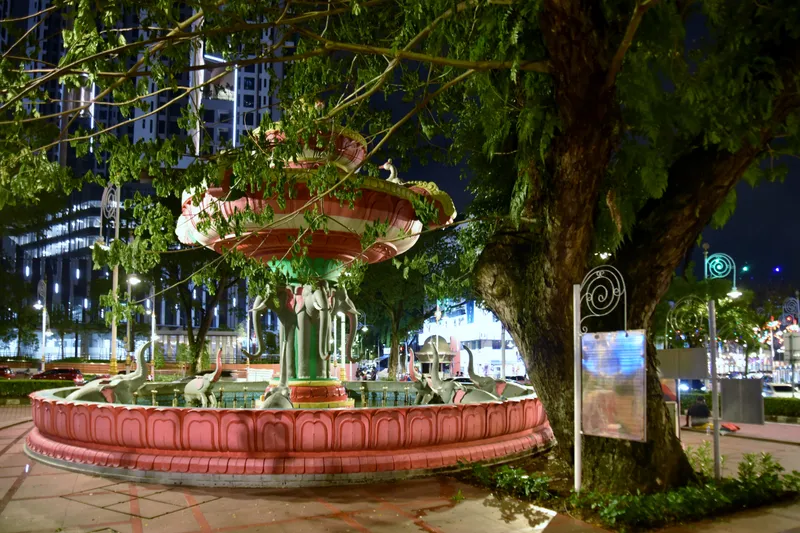 A tiered pink fountain on a base of carved elephants, lit at night under a large tree, with an apartment tower behind