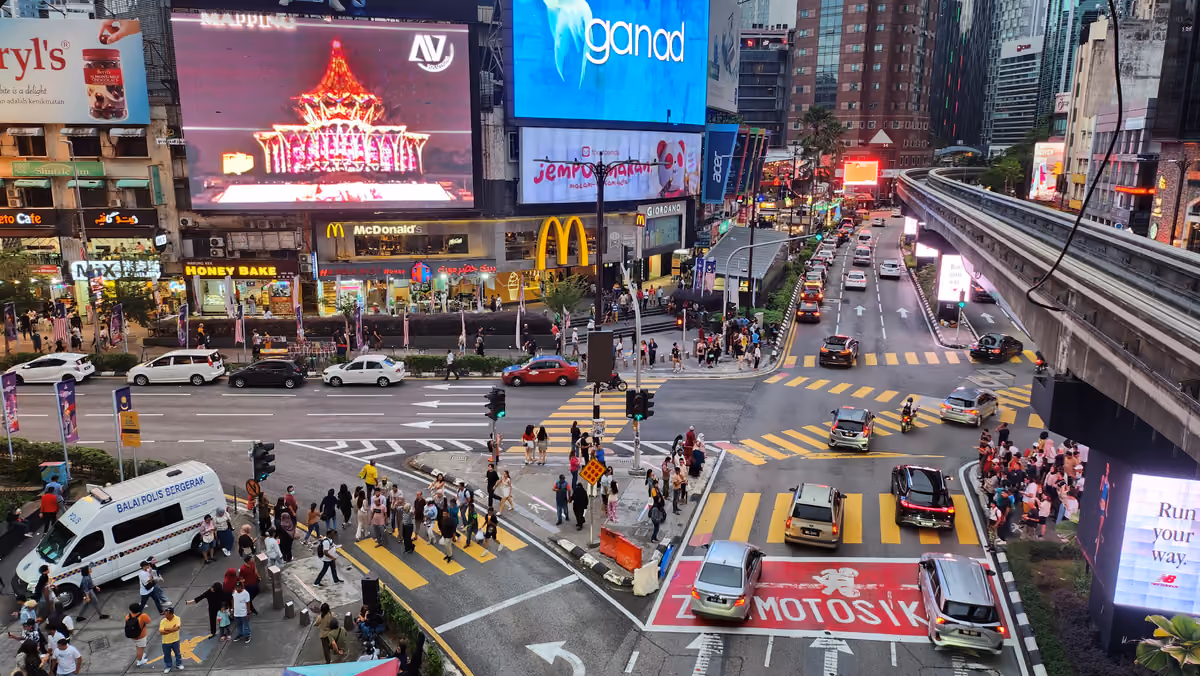 A busy crossroads seen from above in the evening, with people on the zebra crossings, large lit advertising screens on the buildings and the monorail track curving along the right