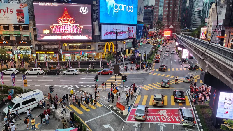 A busy crossroads seen from above in the evening, with people on the zebra crossings, large lit advertising screens on the buildings and the monorail track curving along the right