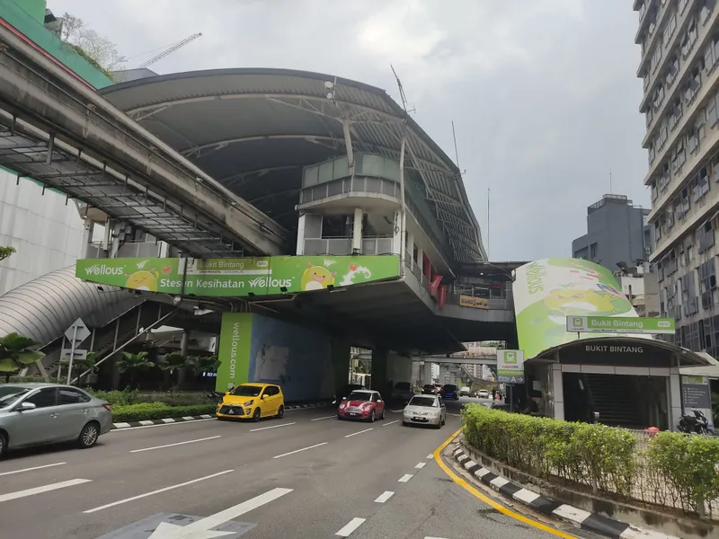 An elevated monorail station with a curved roof above a wide street, with a green Bukit Bintang sign on the station and on a street-level entrance