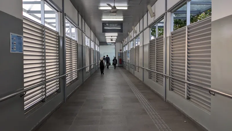 A long covered walkway with louvred walls, a tiled floor and a ceiling fan, with a few people walking away at the far end