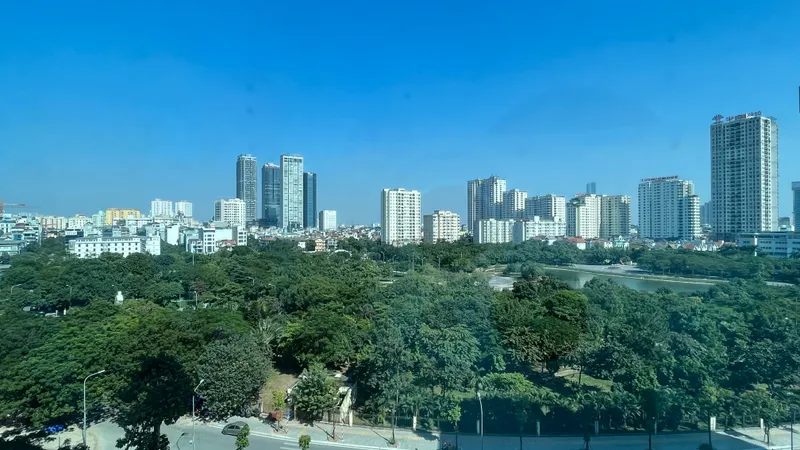 A wide view over the green treetops of Cau Giay Park and a small lake, with white apartment towers under a clear blue sky beyond