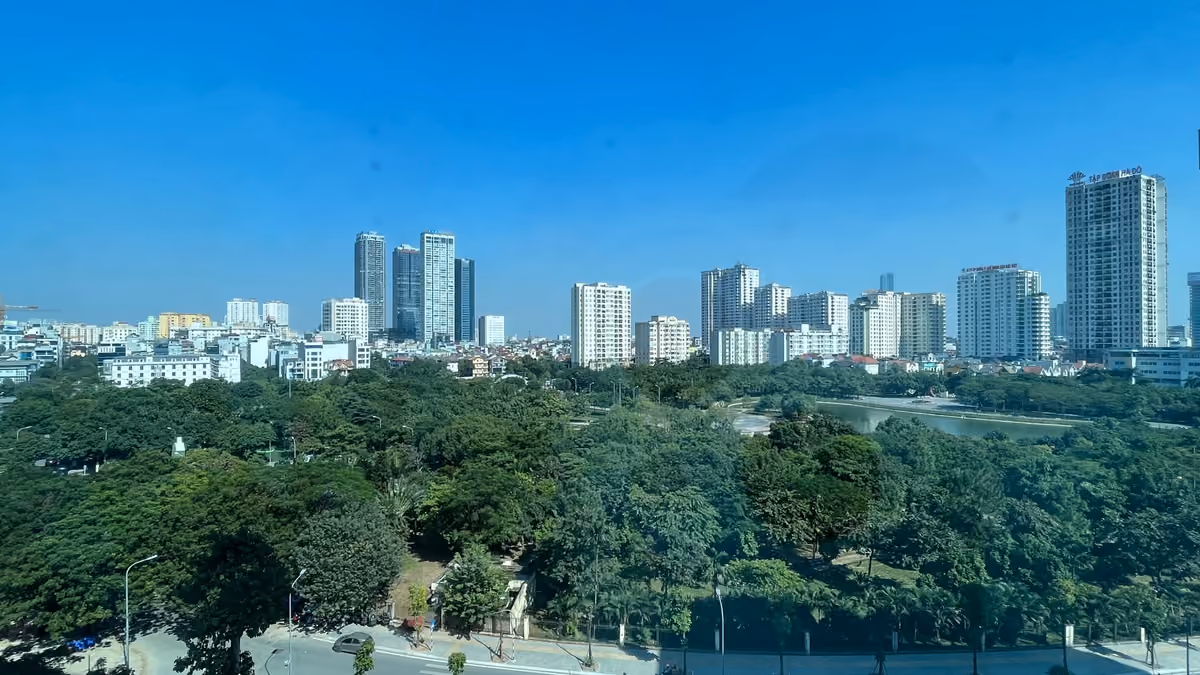 A wide view over the green treetops of Cau Giay Park and a small lake, with white apartment towers under a clear blue sky beyond