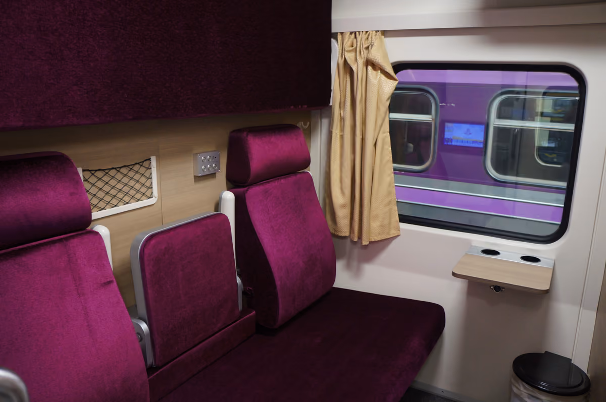 A first-class sleeper cabin with two purple padded seats, a gold curtain at the window and a small fold-down table