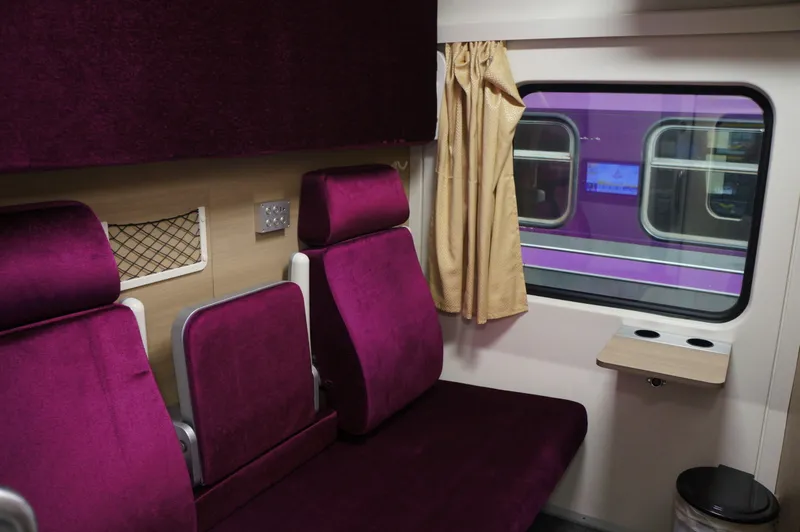 A first-class sleeper cabin with two purple padded seats, a gold curtain at the window and a small fold-down table