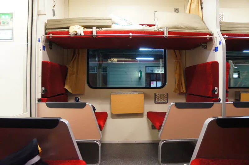 Inside a second-class sleeper car: two red facing seats by a window, with an upper berth folded down above them and a mattress and pillow on it