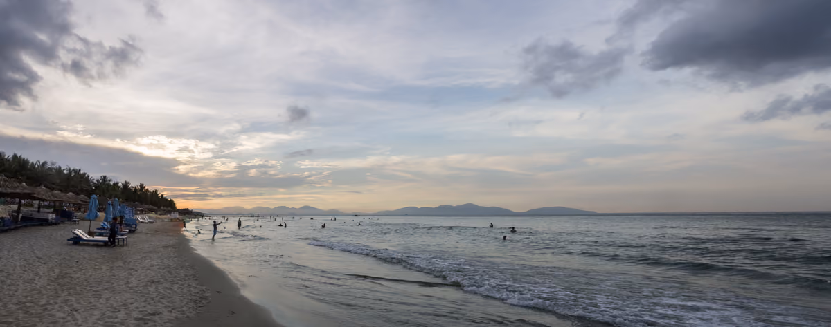 A long beach with thatched sunshades and loungers, a few swimmers in calm water and mountains on the horizon