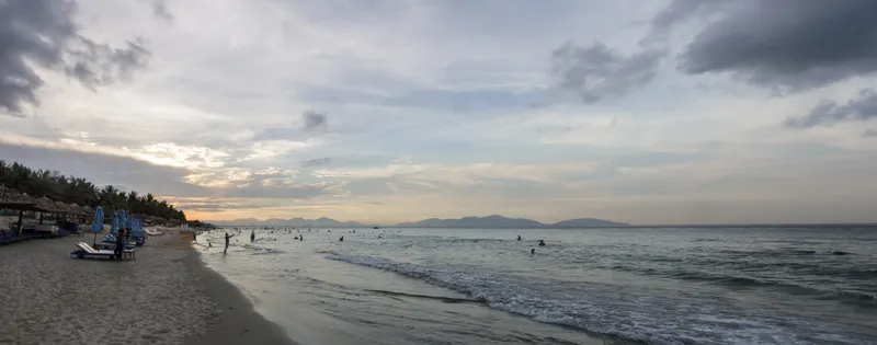 A long beach with thatched sunshades and loungers, a few swimmers in calm water and mountains on the horizon