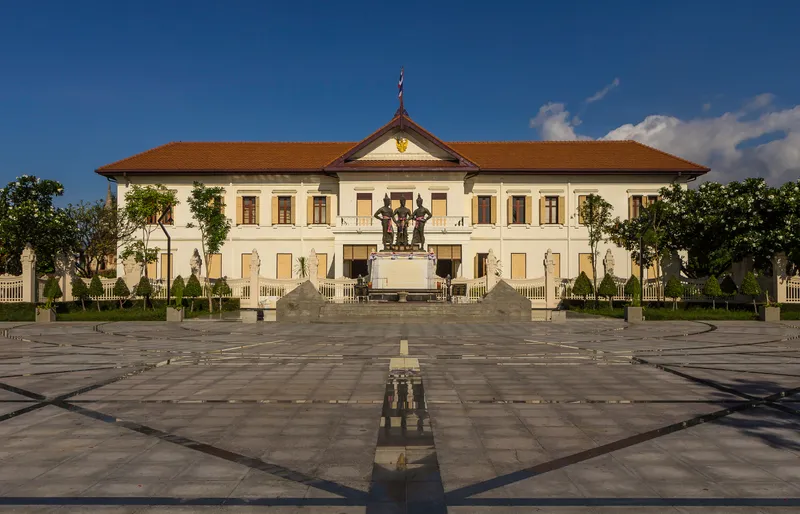 A white two-storey colonial-style building with a red tiled roof, with bronze statues of three standing kings on a plinth in the paved square in front