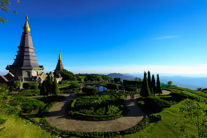 Two tall tiered chedis on a green hilltop above a round formal garden with paths and clipped trees, with blue mountain ridges in the distance