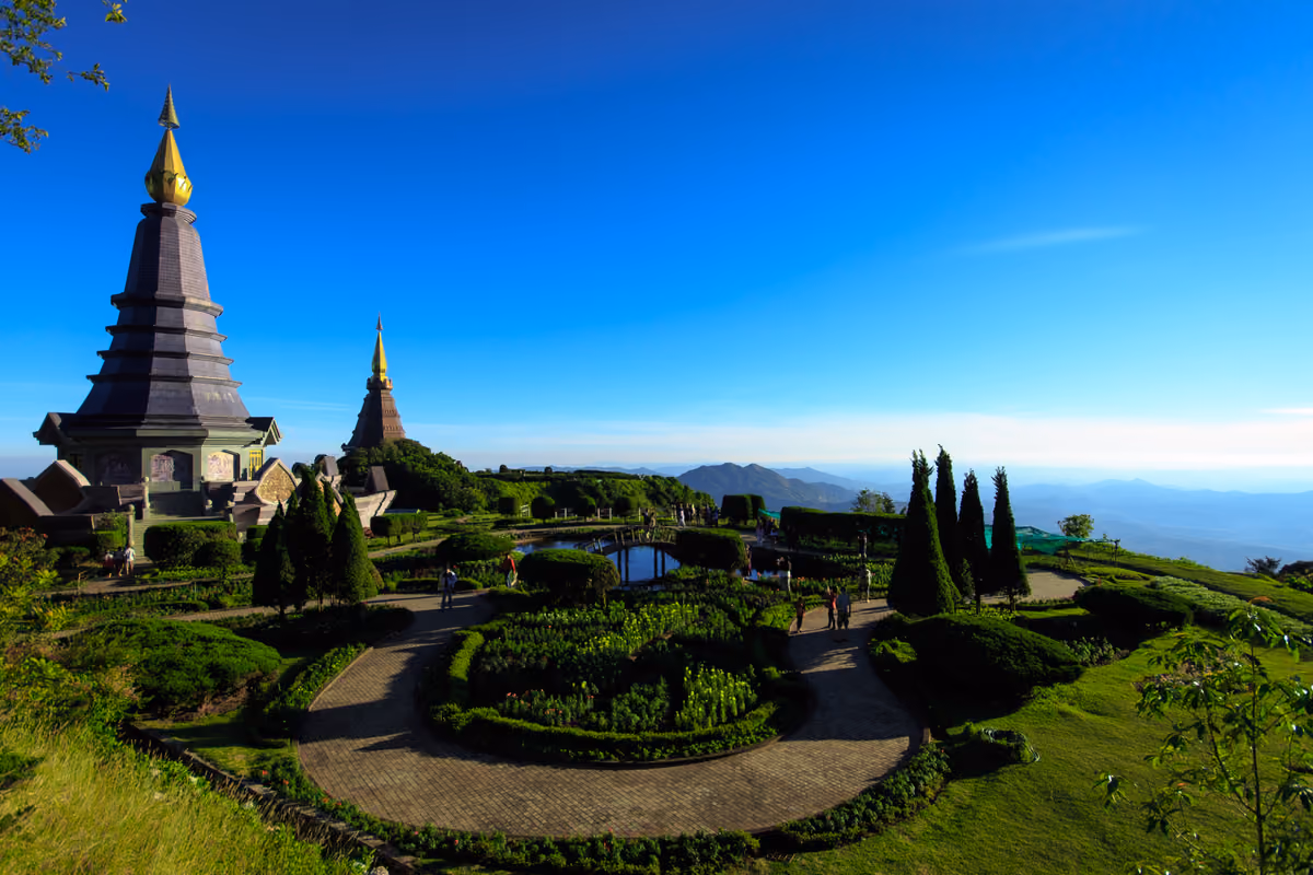 Two tall tiered chedis on a green hilltop above a round formal garden with paths and clipped trees, with blue mountain ridges in the distance