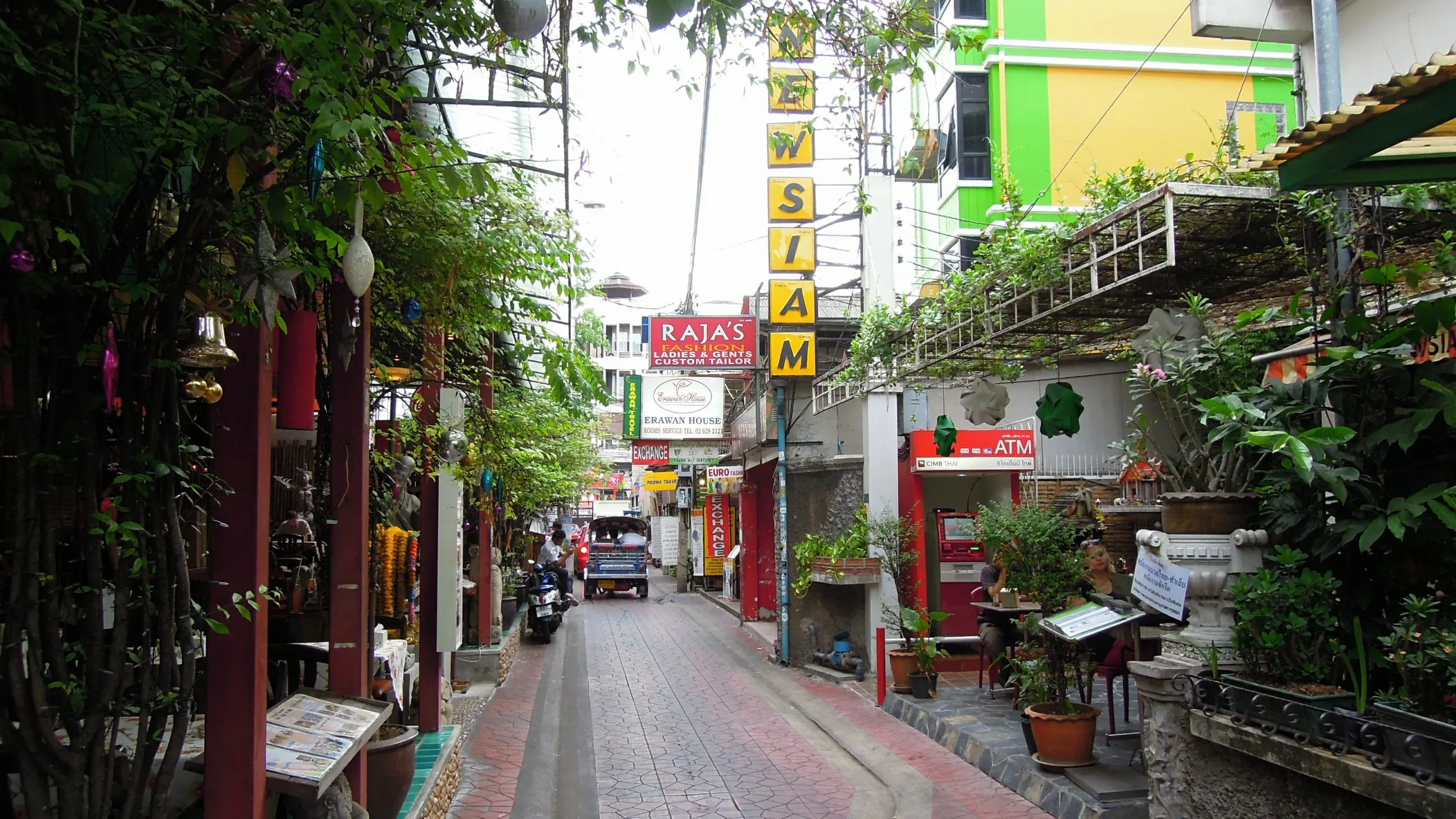 A narrow paved lane lined with plants and guesthouse signs, with a tall yellow vertical sign, a tuk-tuk and a motorbike further down