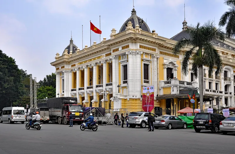 The yellow and white Hanoi Opera House, with columns and domed roofs, and motorbikes and cars on the square in front
