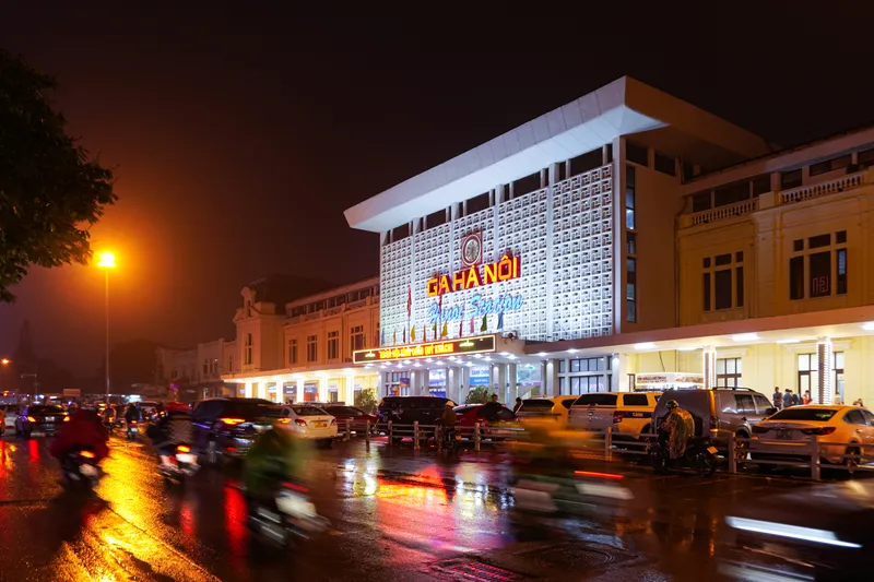 The white, latticed front of Hanoi Station lit up at night, with its Ga Hà Nội sign, taxis parked in front and motorbikes moving along the wet street