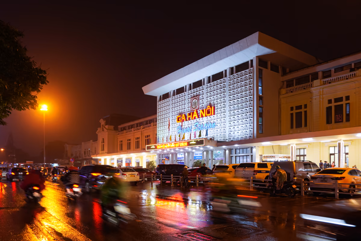 The white, latticed front of Hanoi Station lit up at night, with its Ga Hà Nội sign, taxis parked in front and motorbikes moving along the wet street