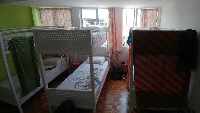 A hostel dorm room with three white metal bunk beds, curtains on some bunks, a parquet floor and windows at the back