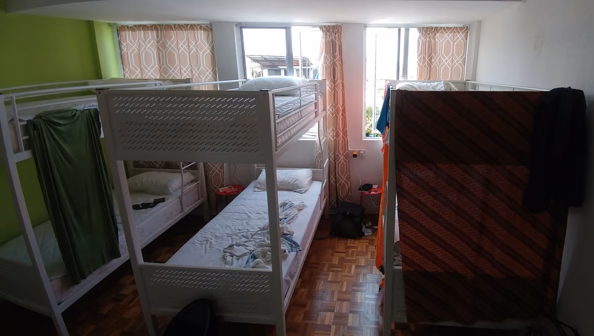 A hostel dorm room with three white metal bunk beds, curtains on some bunks, a parquet floor and windows at the back
