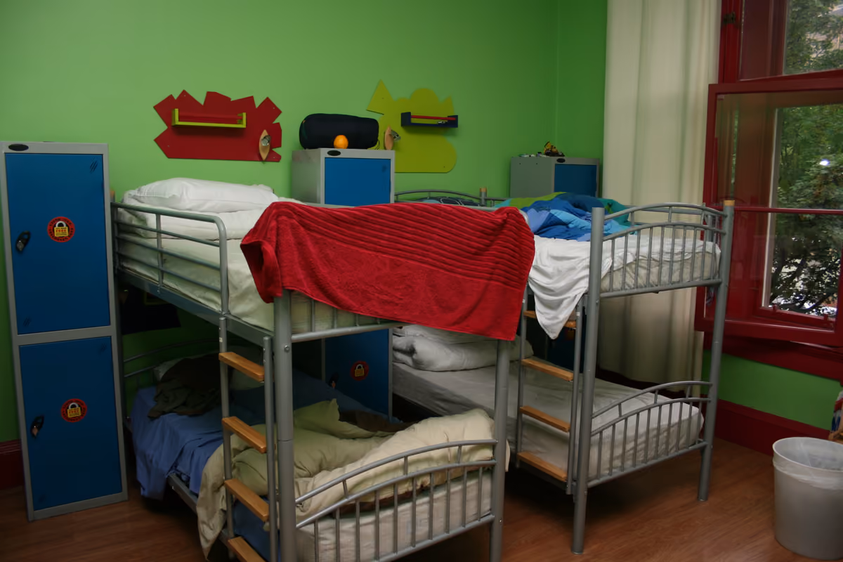 A hostel dorm with metal bunk beds and tall blue lockers, each marked with a padlock symbol, against a green wall