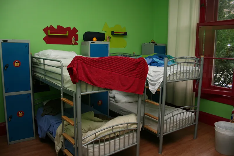A hostel dorm with metal bunk beds and tall blue lockers, each marked with a padlock symbol, against a green wall