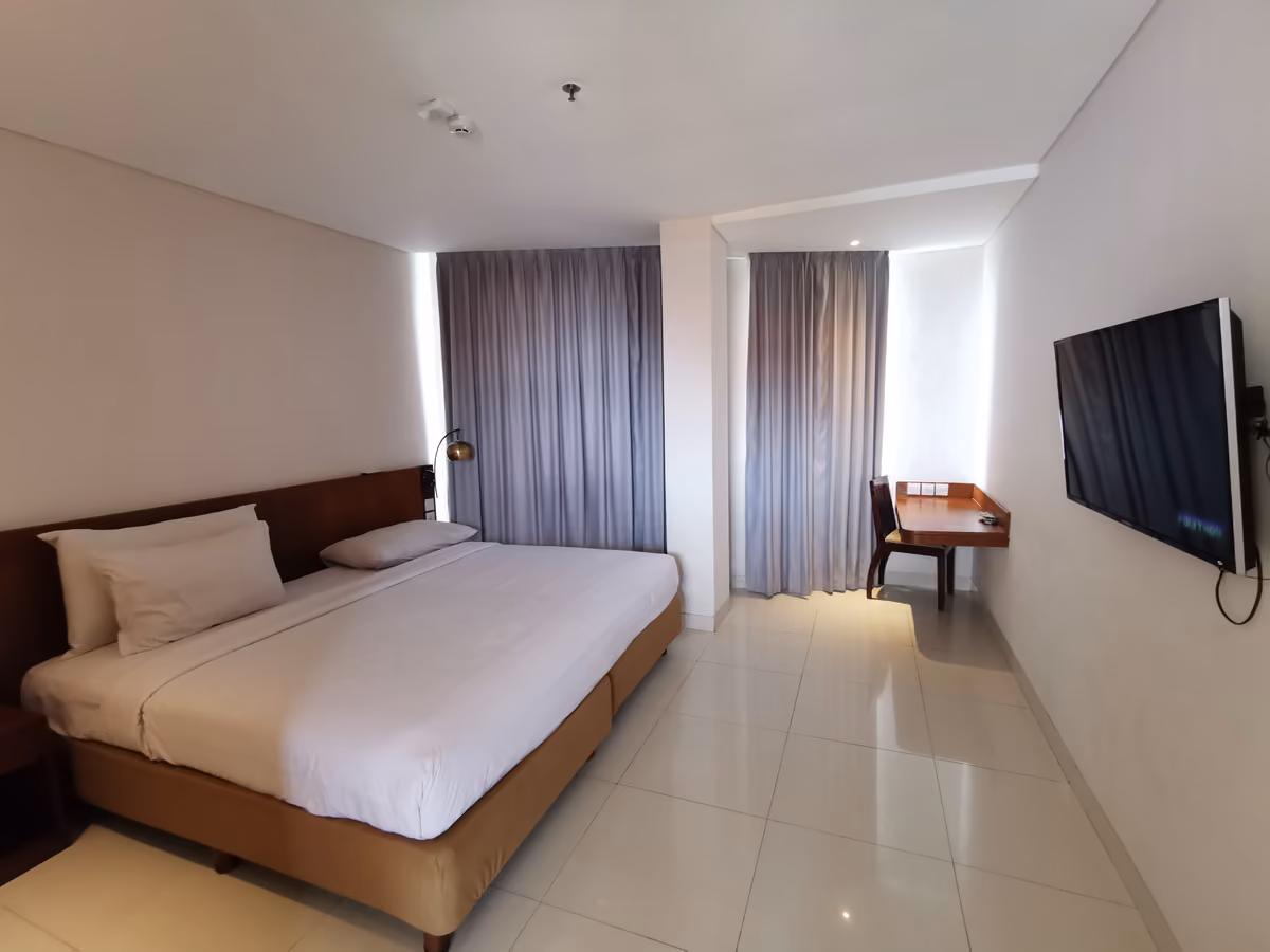 A hotel double room with a large white bed, grey curtains, a small desk and chair by the window and a television on the wall
