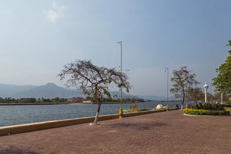 A wide brick riverside promenade in Kampot with a young tree and lamp posts, the river and low green mountains under a hazy blue sky
