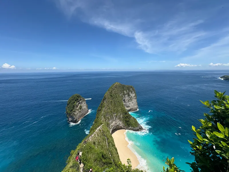 A green, steep-sided headland shaped like a dinosaur’s head dropping to a curved white beach and turquoise sea under a clear sky