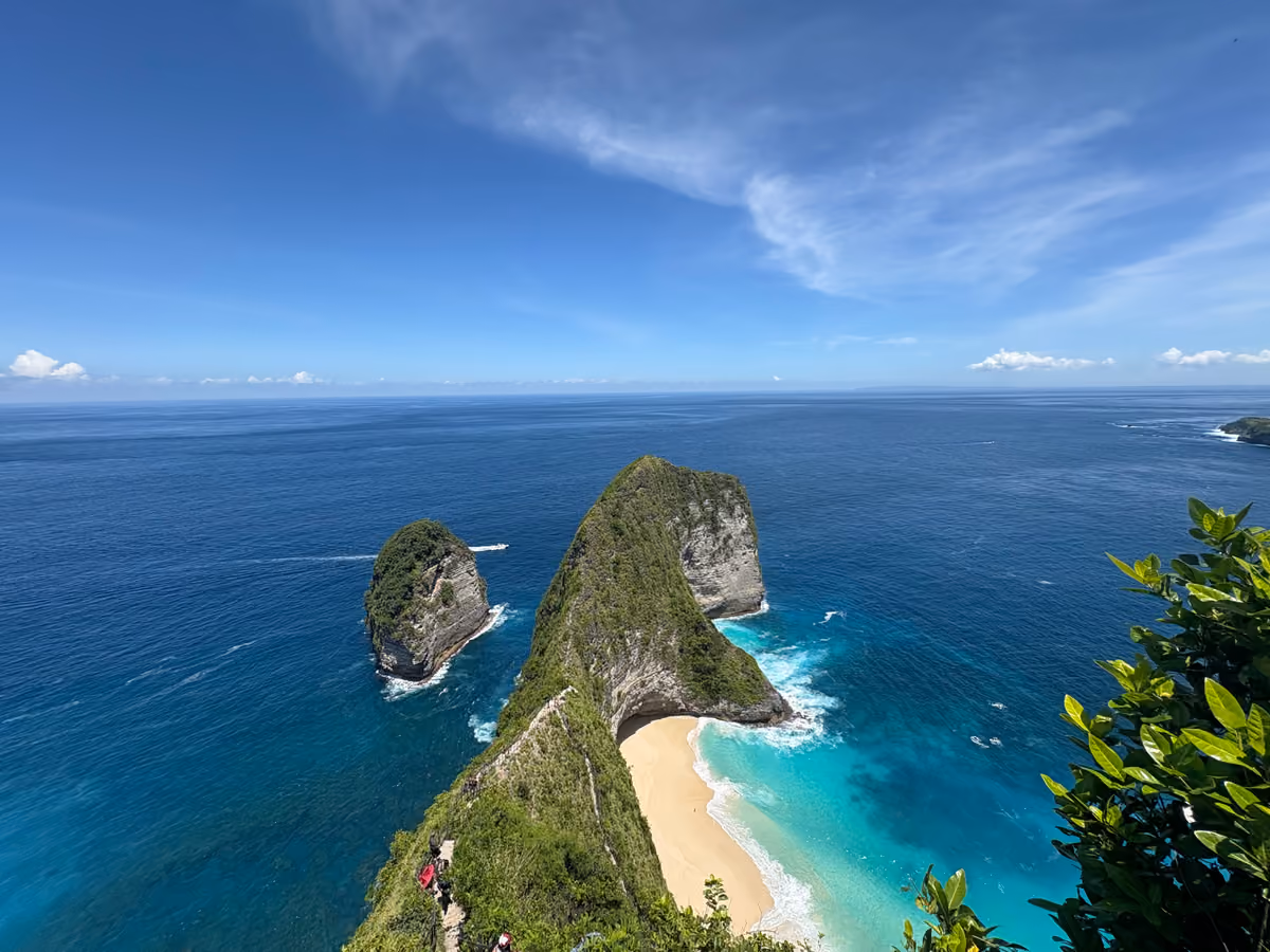 A green, steep-sided headland shaped like a dinosaur’s head dropping to a curved white beach and turquoise sea under a clear sky