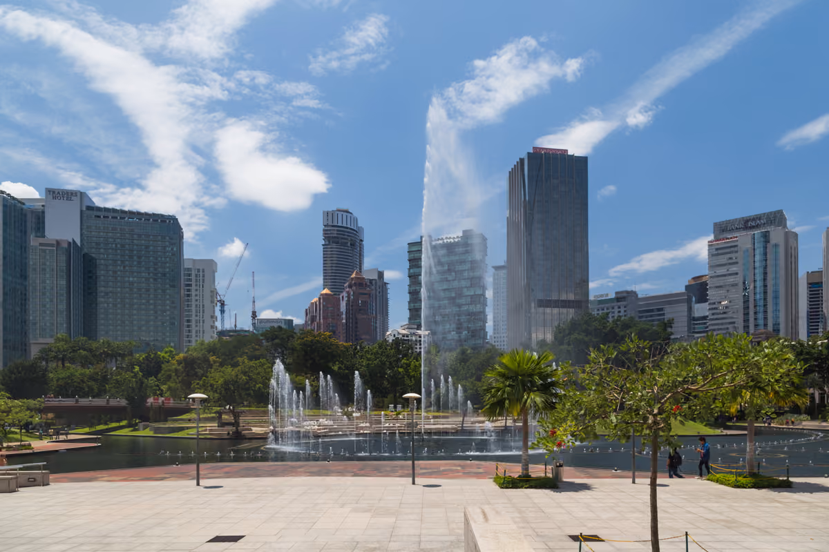 A paved terrace above a lake in a city park, with fountains, palm trees and green lawns, and tall office towers behind under a blue sky