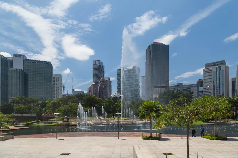 A paved terrace above a lake in a city park, with fountains, palm trees and green lawns, and tall office towers behind under a blue sky