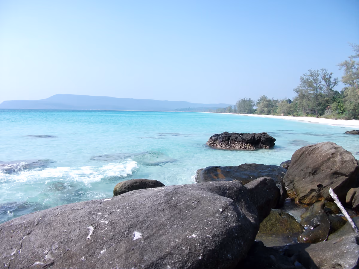 A long white-sand beach curving away along turquoise water, with dark boulders in the foreground and wooded hills on the horizon
