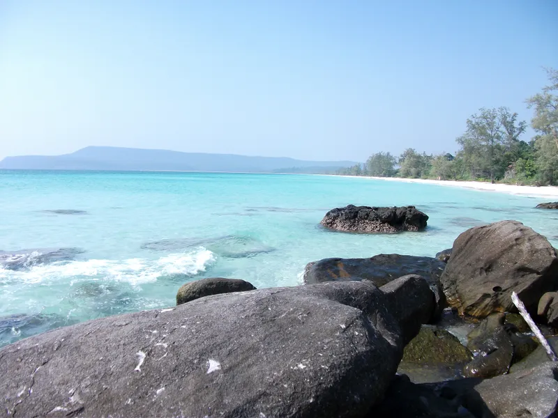 A long white-sand beach curving away along turquoise water, with dark boulders in the foreground and wooded hills on the horizon