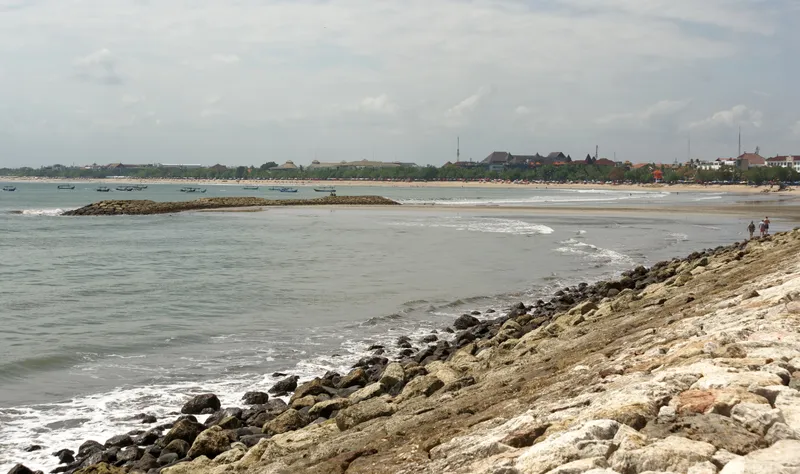 A wide, curving beach at Kuta seen from a rocky sea wall, with a low stone breakwater, small boats offshore and trees and low buildings along the shore under a pale, cloudy sky