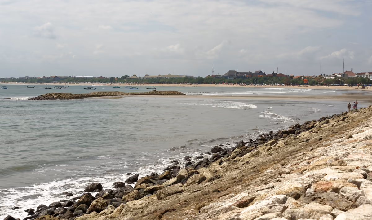 A wide, curving beach at Kuta seen from a rocky sea wall, with a low stone breakwater, small boats offshore and trees and low buildings along the shore under a pale, cloudy sky