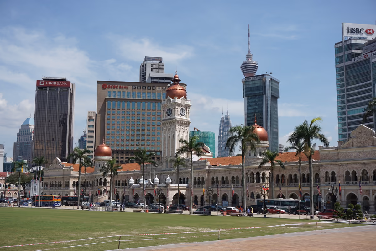 A wide lawn in front of a long arcaded building with a clock tower and copper domes, office towers and the Petronas Twin Towers behind