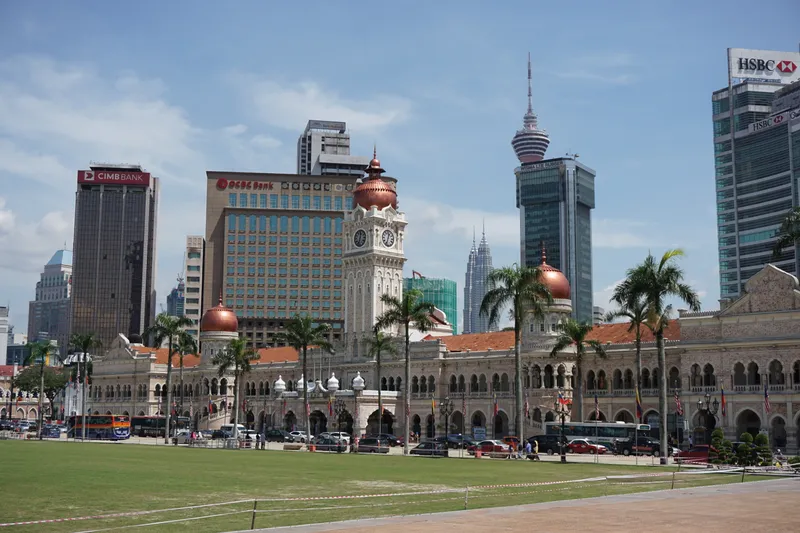 A wide lawn in front of a long arcaded building with a clock tower and copper domes, office towers and the Petronas Twin Towers behind