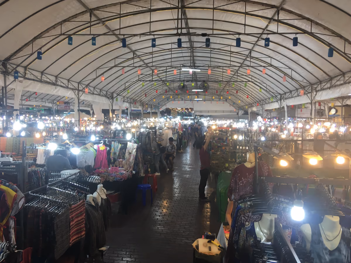 A long covered market hall at night, lit by bare bulbs and paper lanterns, with clothes and souvenirs on racks on both sides of a brick walkway