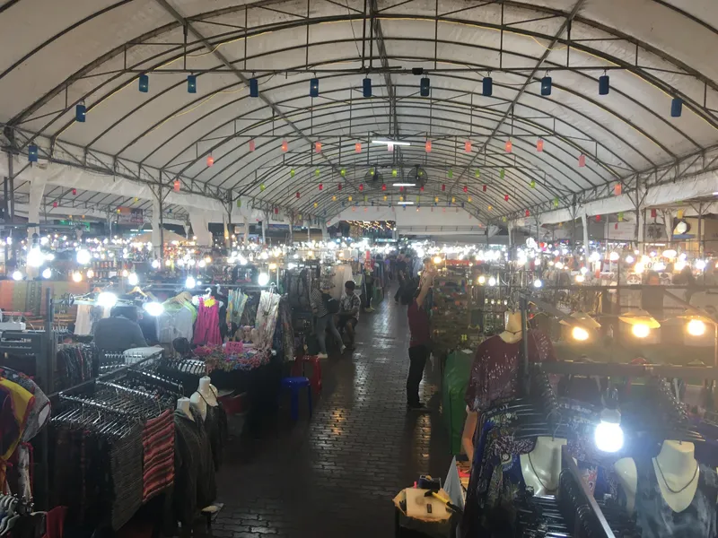 A long covered market hall at night, lit by bare bulbs and paper lanterns, with clothes and souvenirs on racks on both sides of a brick walkway