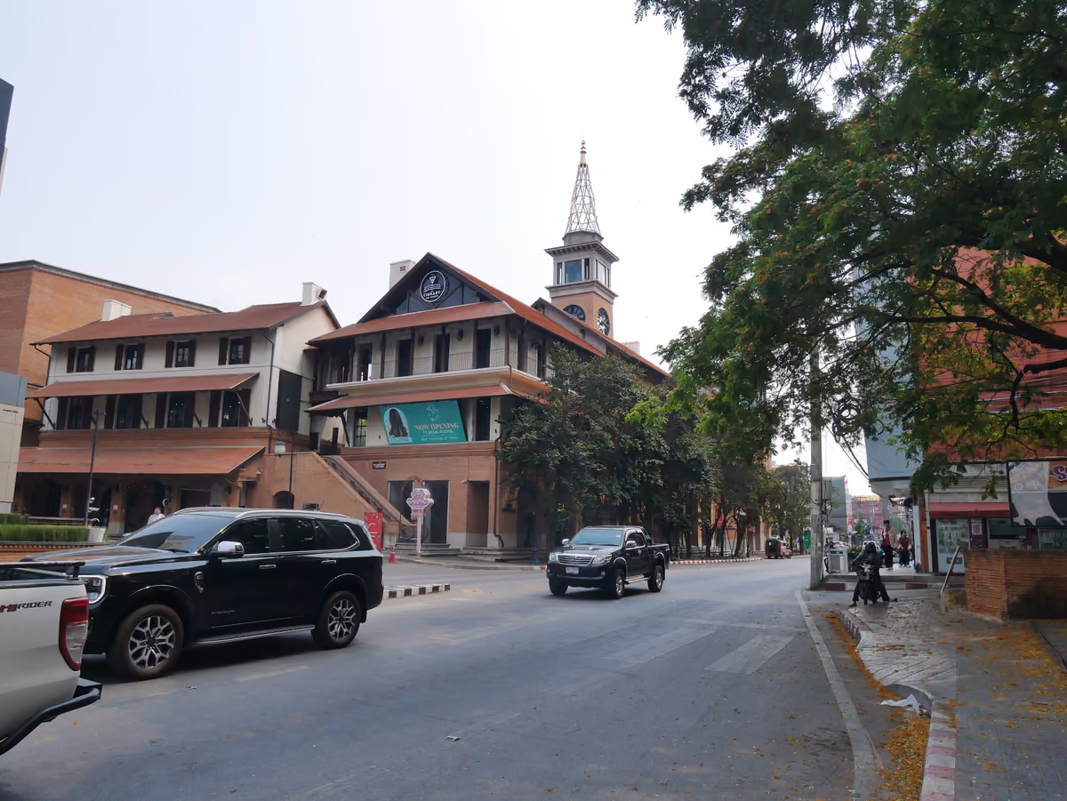 A street of brick buildings with a tall clock tower, cars driving past and trees along the pavement on the right