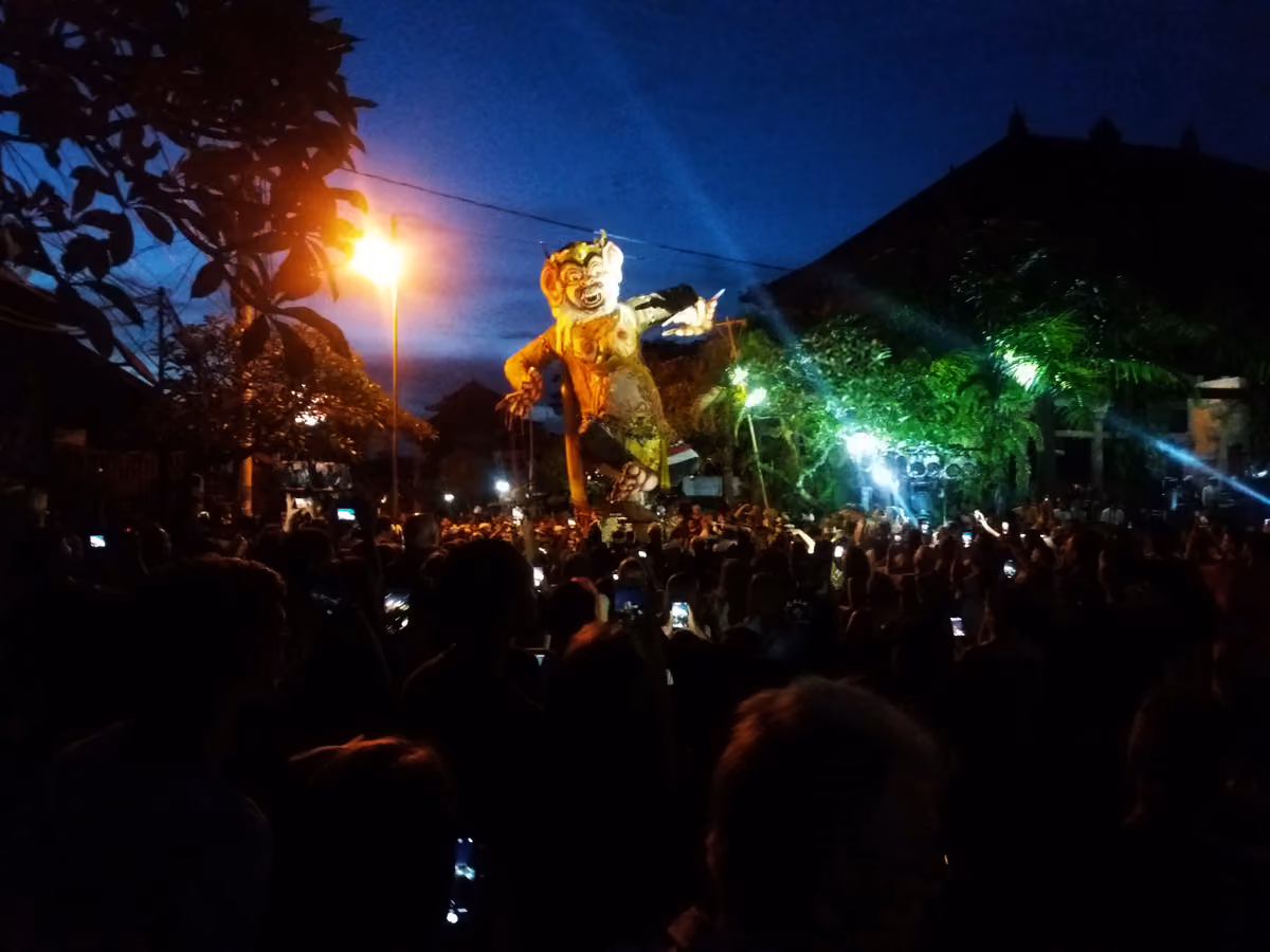 A tall, painted ogoh-ogoh demon figure lit by streetlights at dusk above a dark crowd holding up phones