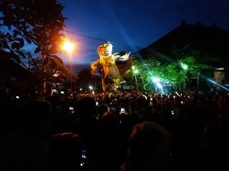 A tall, painted ogoh-ogoh demon figure lit by streetlights at dusk above a dark crowd holding up phones