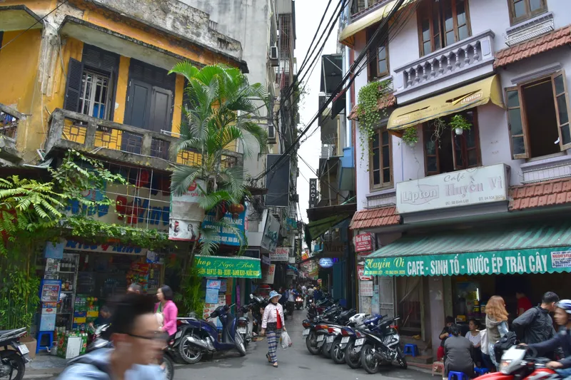 A narrow Old Quarter street of tall, narrow shopfront houses with balconies, shop signs and parked motorbikes