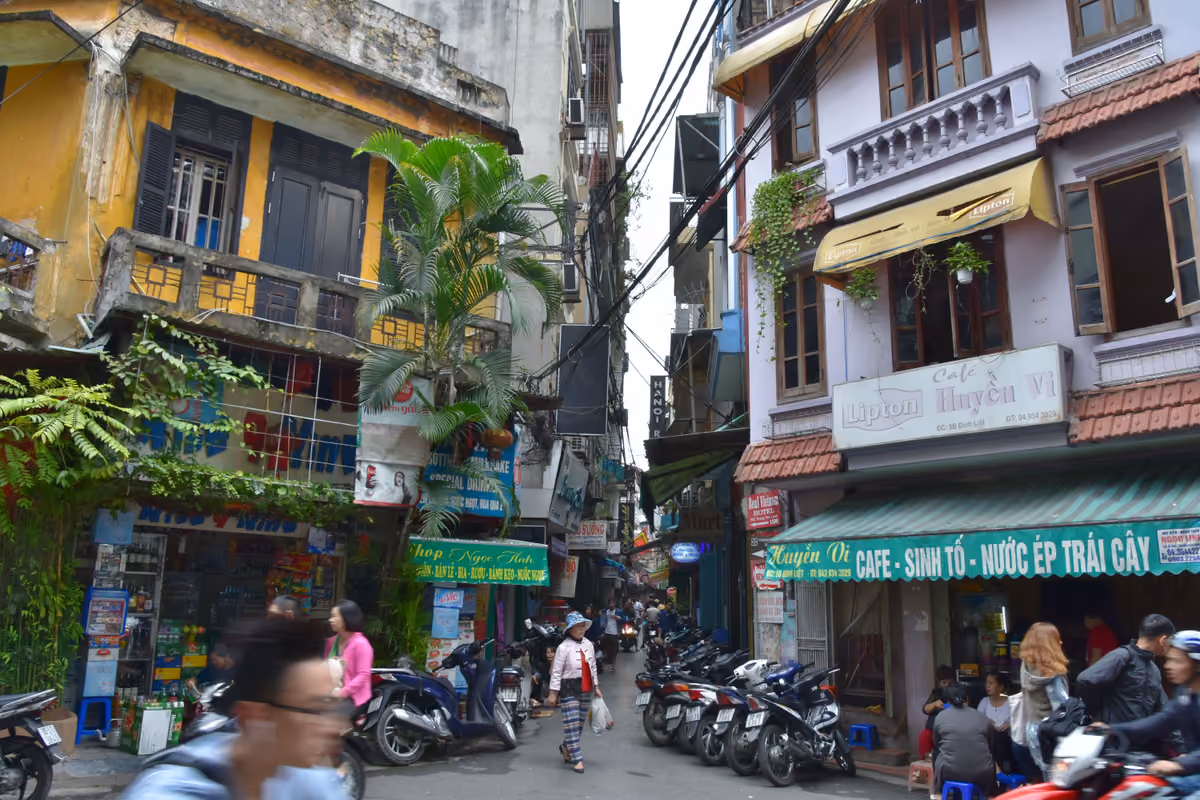 A narrow Old Quarter street of tall, narrow shopfront houses with balconies, shop signs and parked motorbikes